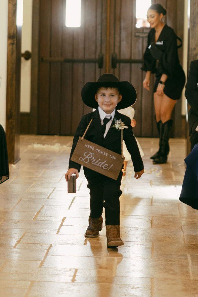 little boy walking down the aisle with a sign around his neck that says here comes the bride