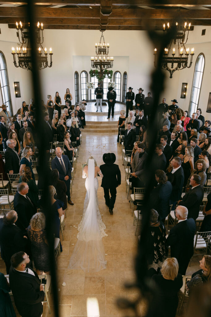 bride walking down the aisle with her father before she gets married, holding a big bridal bouquet and her father is wearing all black with a black cowboy hat