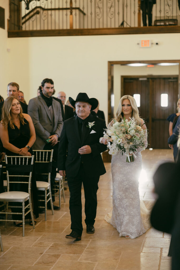 bride walking down the aisle with her father before she gets married, holding a big bridal bouquet and her father is wearing all black with a black cowboy hat