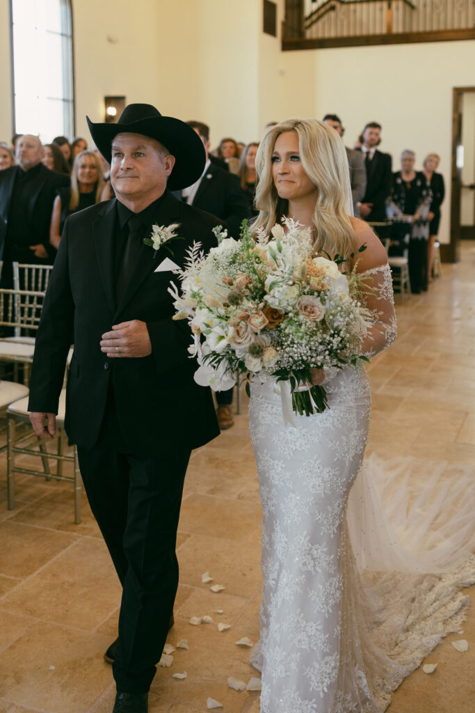 bride walking down the aisle with her father before she gets married, holding a big bridal bouquet and her father is wearing all black with a black cowboy hat