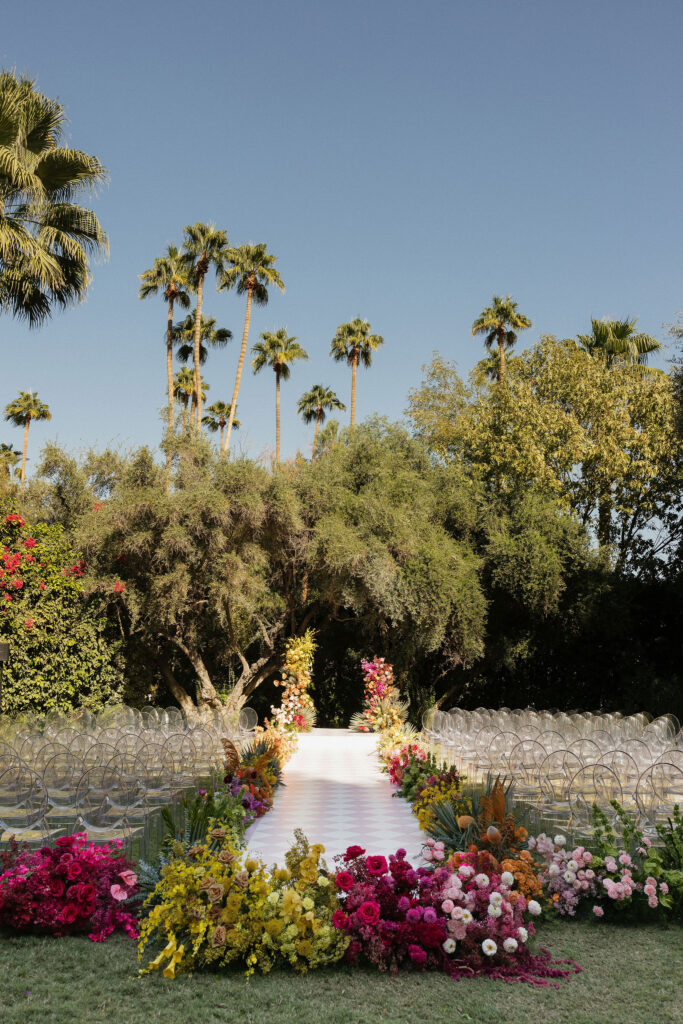 Wedding aisle with floral decorations - A picturesque wedding aisle lined with colorful flowers in pinks, reds, and yellows leads to an altar framed by greenery and tall palms in the background.