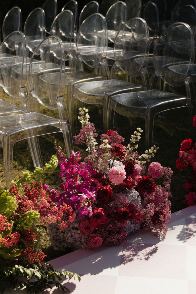 Ghost chairs with floral arrangements - Transparent "ghost" chairs arranged for the ceremony, with a lush arrangement of pink, red, and purple flowers decorating the aisle.