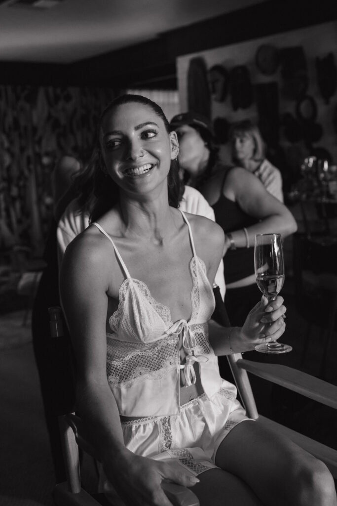 A bride laughs while holding a champagne flute, dressed in white lingerie, seated in a relaxed setting as she prepares for her wedding day.