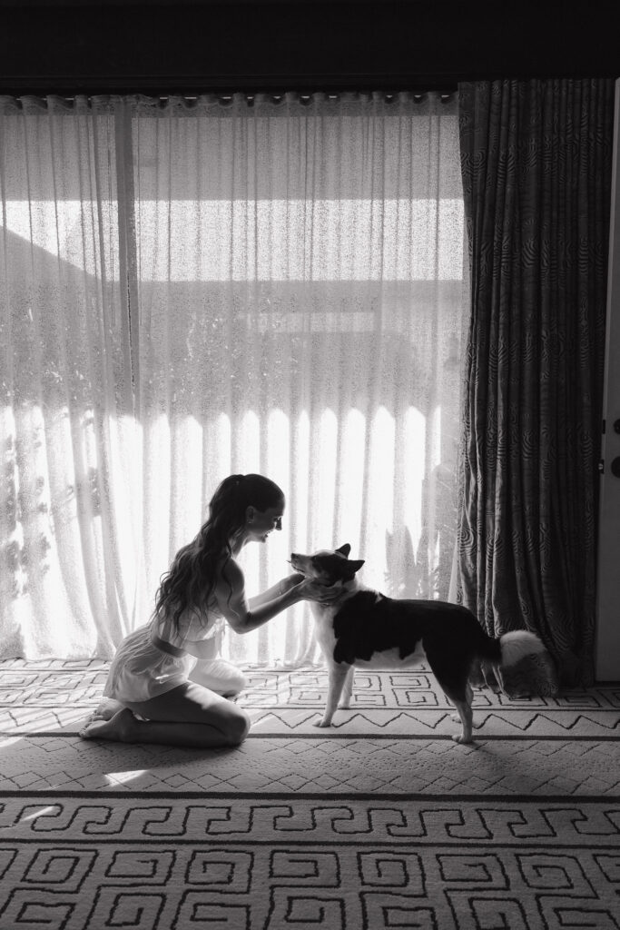 Bride kneeling with dog - A black-and-white photo of the bride kneeling and smiling at her dog in a beautifully lit room, with light streaming through sheer curtains.