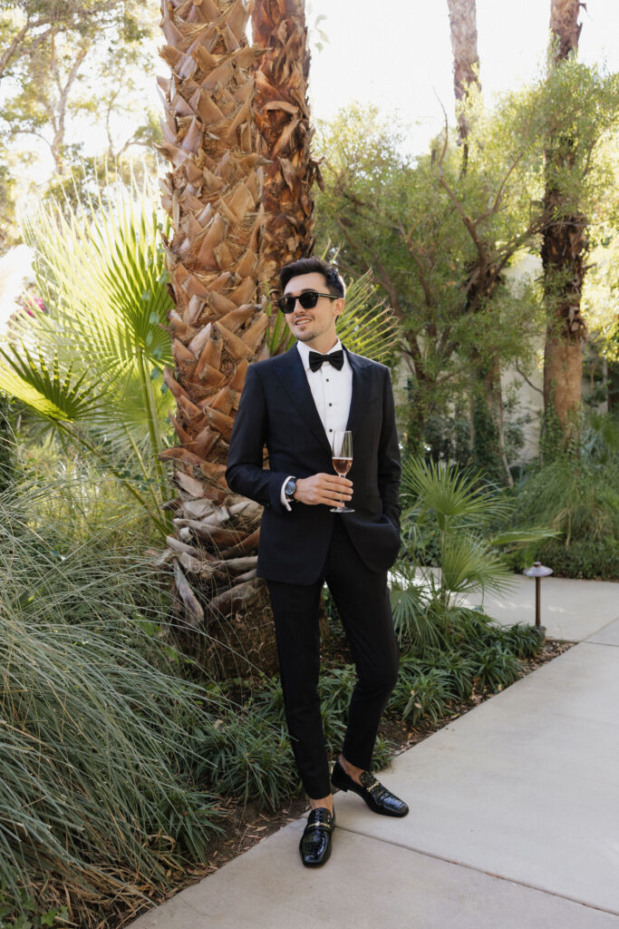 The groom stands confidently in a black tuxedo, holding a glass of champagne with sunglasses on, posing against the backdrop of towering palm trees at Park Palm Springs Hotel.