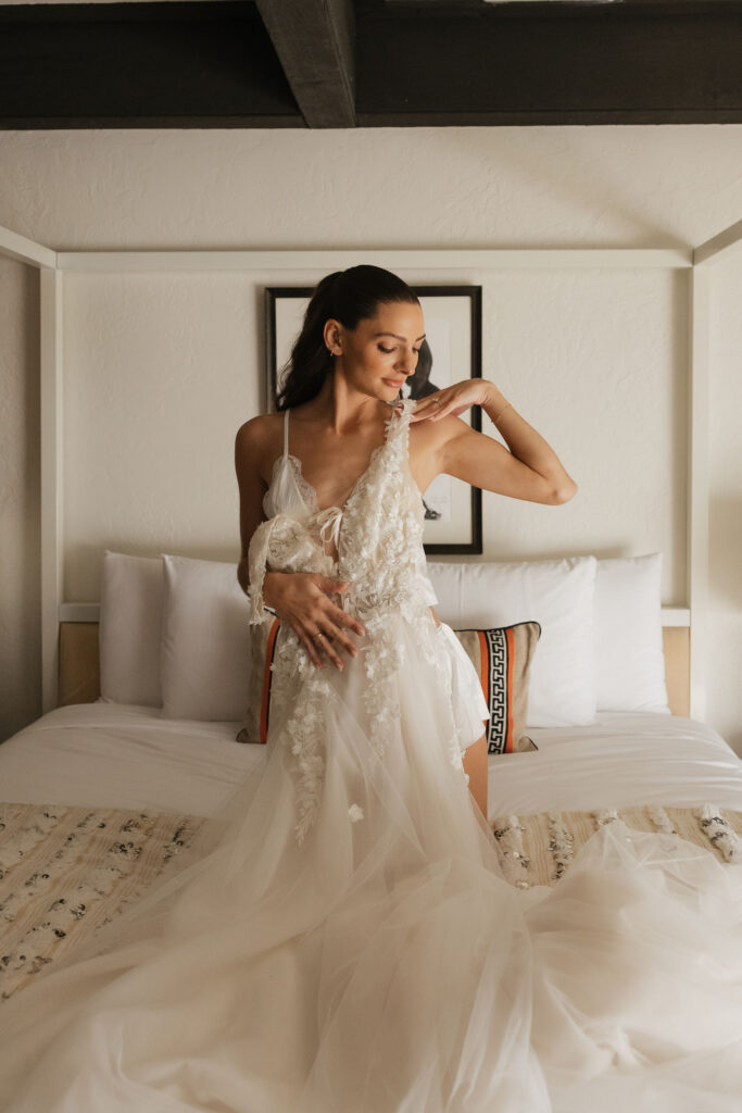 The bride adjusts the strap of her intricately detailed wedding dress while standing on a bed in a bright, cozy room at Park Palm Springs Hotel, capturing a serene pre-wedding moment.