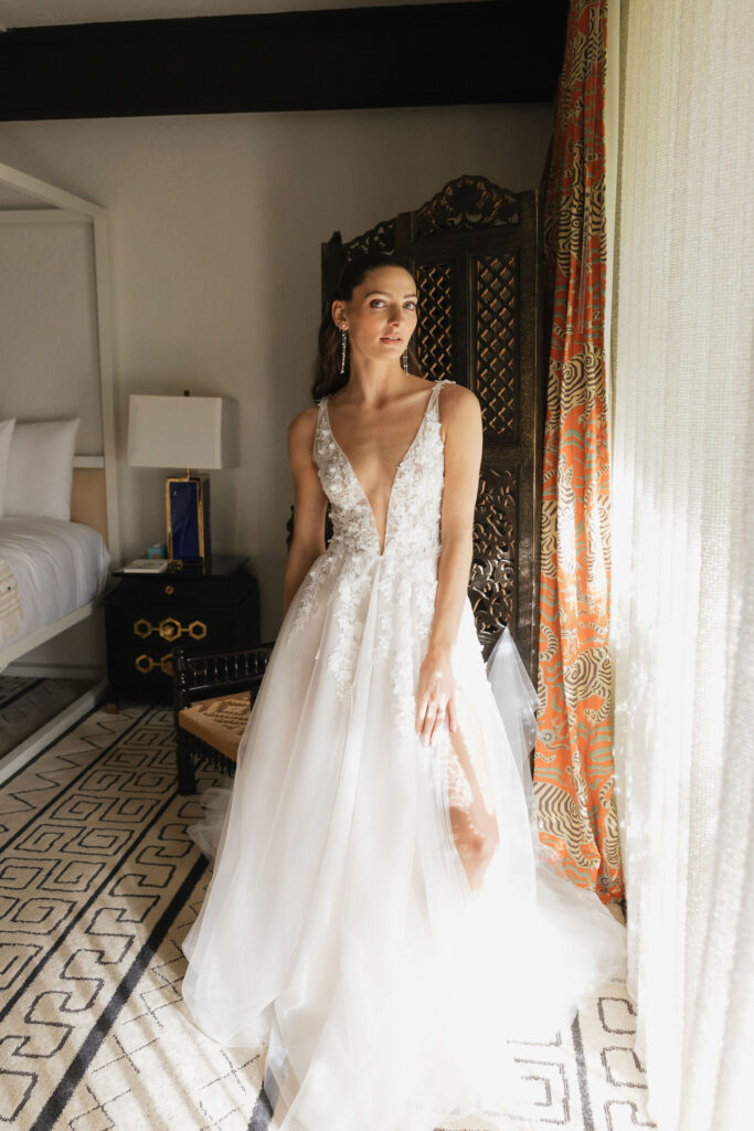 Bride posing in a room lit by natural light, wearing a detailed lace gown with a deep neckline.