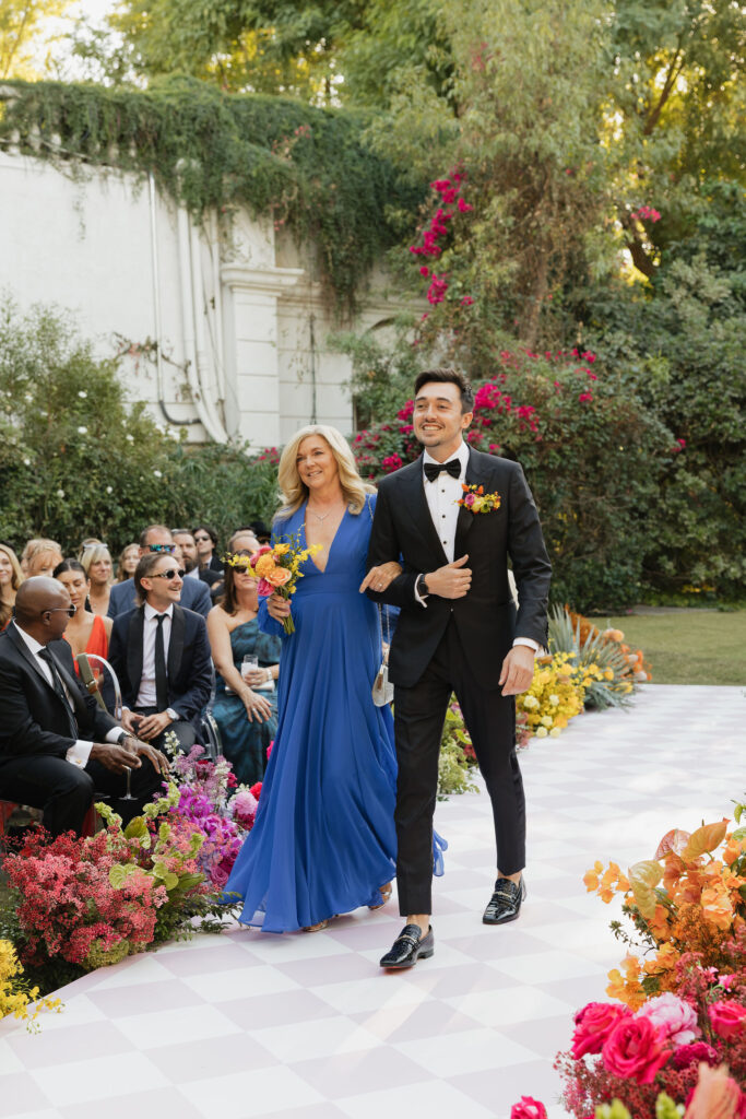 The groom walking down the aisle with his mother at Park Palm Springs Hotel, both smiling and holding colorful bouquets, surrounded by guests and vibrant floral arrangements.