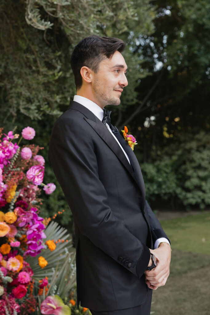 The groom stands with a small, colorful boutonniere pinned to his black tuxedo, smiling as he waits for his bride during the ceremony at Park Palm Springs Hotel.