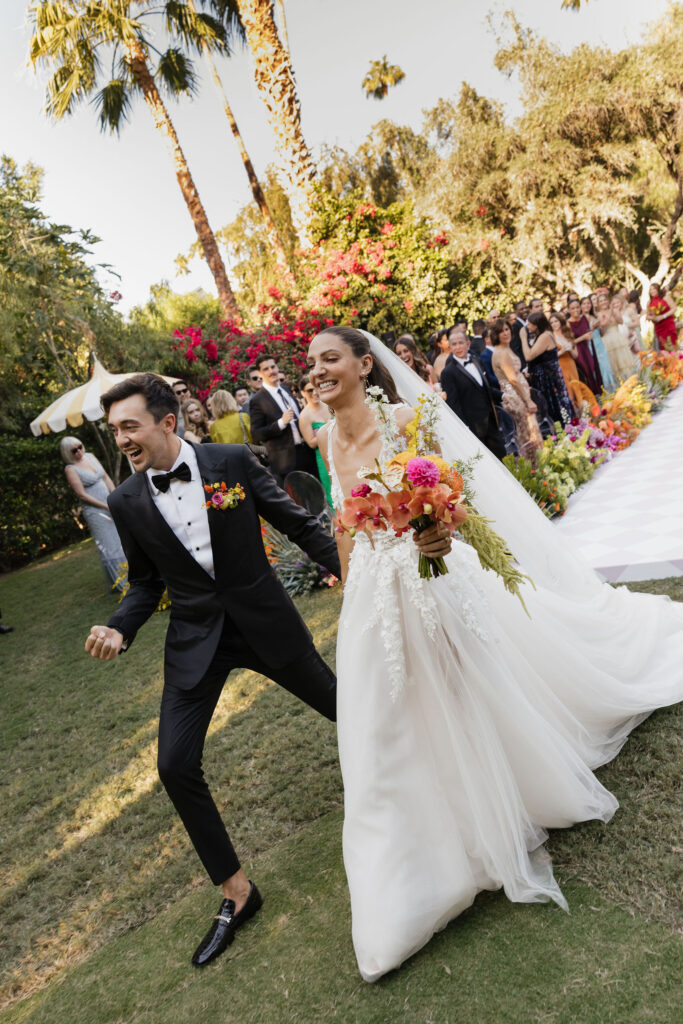 A smiling bride and groom walk down the aisle, surrounded by cheering guests in a lush garden at Park Palm Springs Hotel. The bride holds a colorful bouquet, and both exude happiness as they celebrate their wedding day.