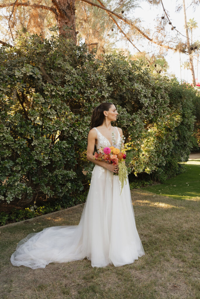 The bride standing outdoors at Park Palm Springs Hotel, holding a colorful bouquet and looking off to the side, with her long veil flowing gently behind her.