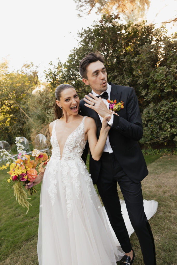 The newlyweds enthusiastically display their wedding rings while smiling and holding hands in a garden setting.