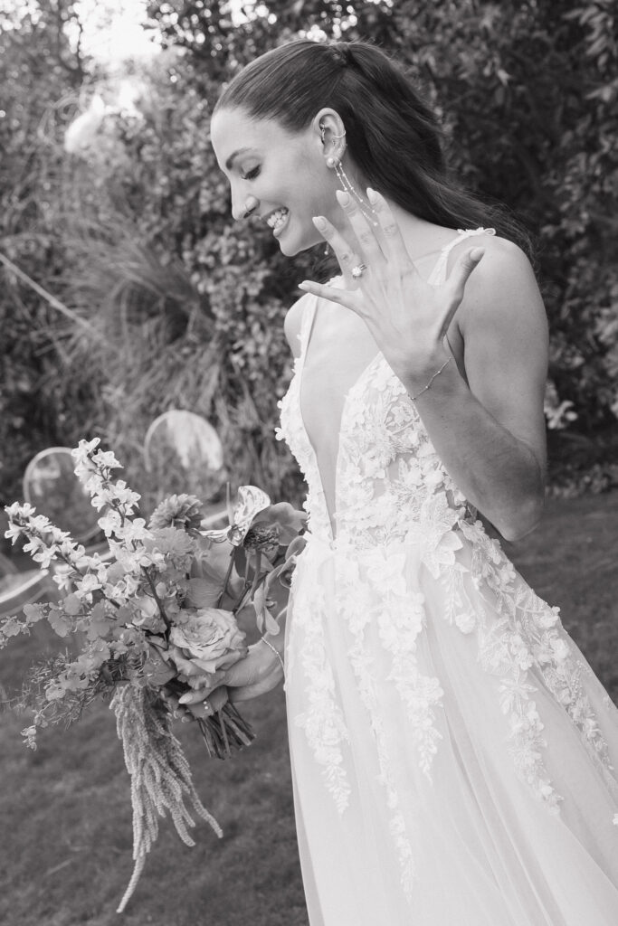 A joyful bride at Park Palm Springs Hotel, holding her bouquet and showing off her wedding ring, with a radiant smile and a lush garden backdrop.