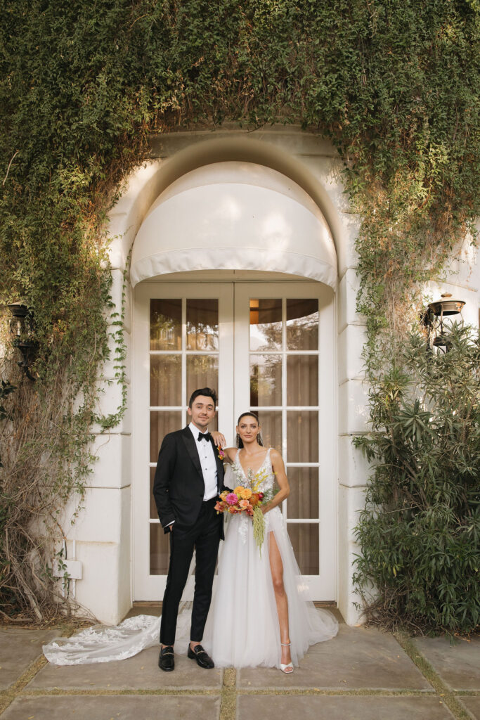 The bride and groom stand together in front of an ivy-covered arch at Park Palm Springs Hotel, posing for a formal wedding portrait.