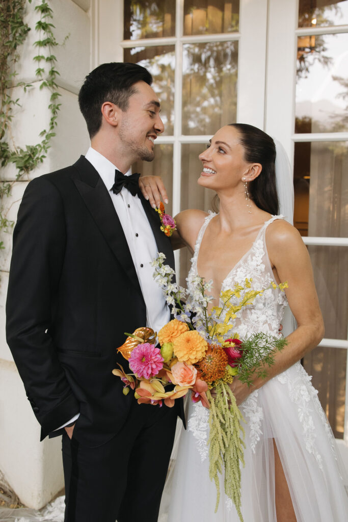 The bride and groom stand in front of white French doors, smiling and gazing at each other, with the bride holding a colorful bouquet.