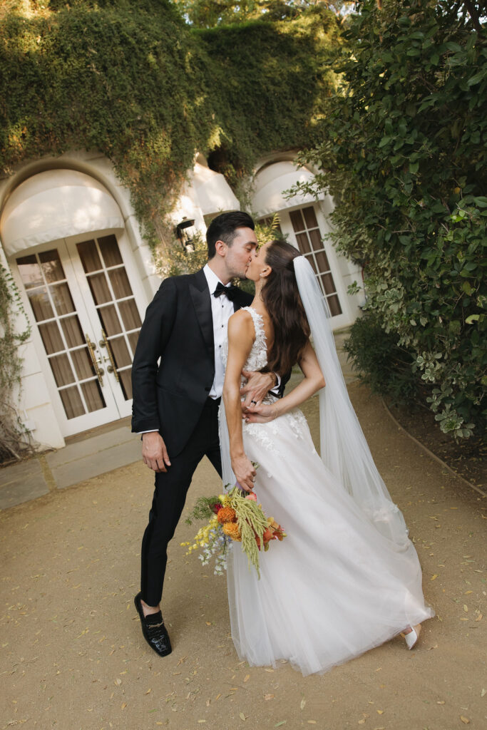 Bride and groom share a kiss in front of a lush, vine-covered doorway, with the bride holding a colorful bouquet.