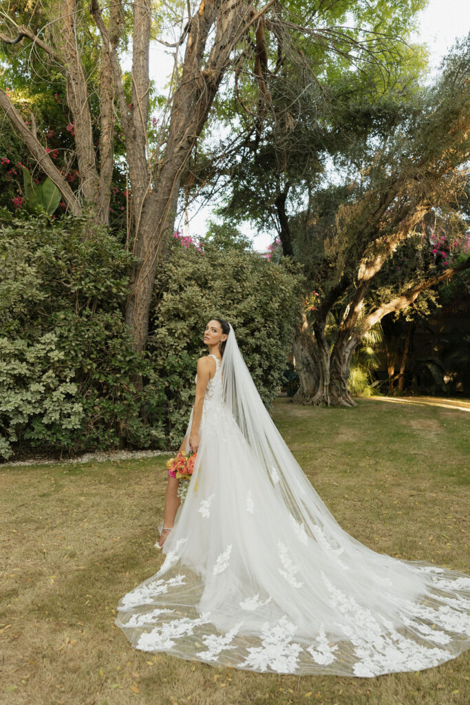 Bride with veil flowing - The bride, holding her bouquet, stands in the garden with her long veil and train flowing behind her, looking over her shoulder.