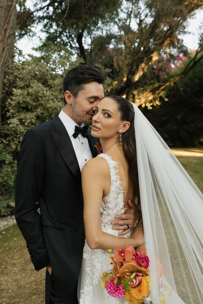 Bride and groom embracing in garden - The groom whispers affectionately into the bride's ear as they stand in a lush garden, the bride holding a vibrant bouquet.