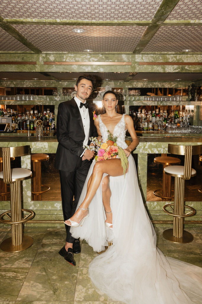 The bride and groom pose together at an elegant, green-marble bar at Park Palm Springs Hotel, the bride holding a bouquet of brightly colored flowers.