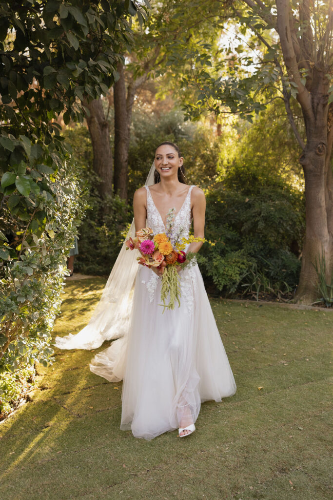 The bride, in a flowy lace wedding gown with floral details, walks down a garden path at Park Palm Springs Hotel, holding a vibrant bouquet of pink, orange, and yellow flowers, with a serene expression on her face.