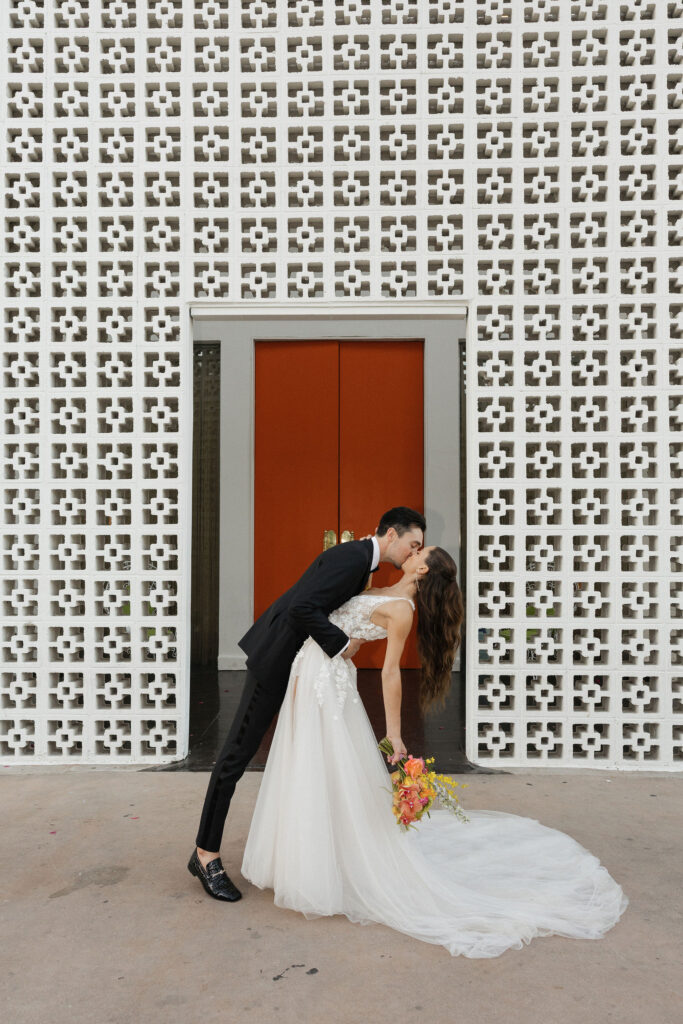 Bride and groom sharing a romantic dip kiss in front of a white patterned wall - The groom dips the bride for a kiss, set against the backdrop of a retro-style patterned wall and bold orange doors.