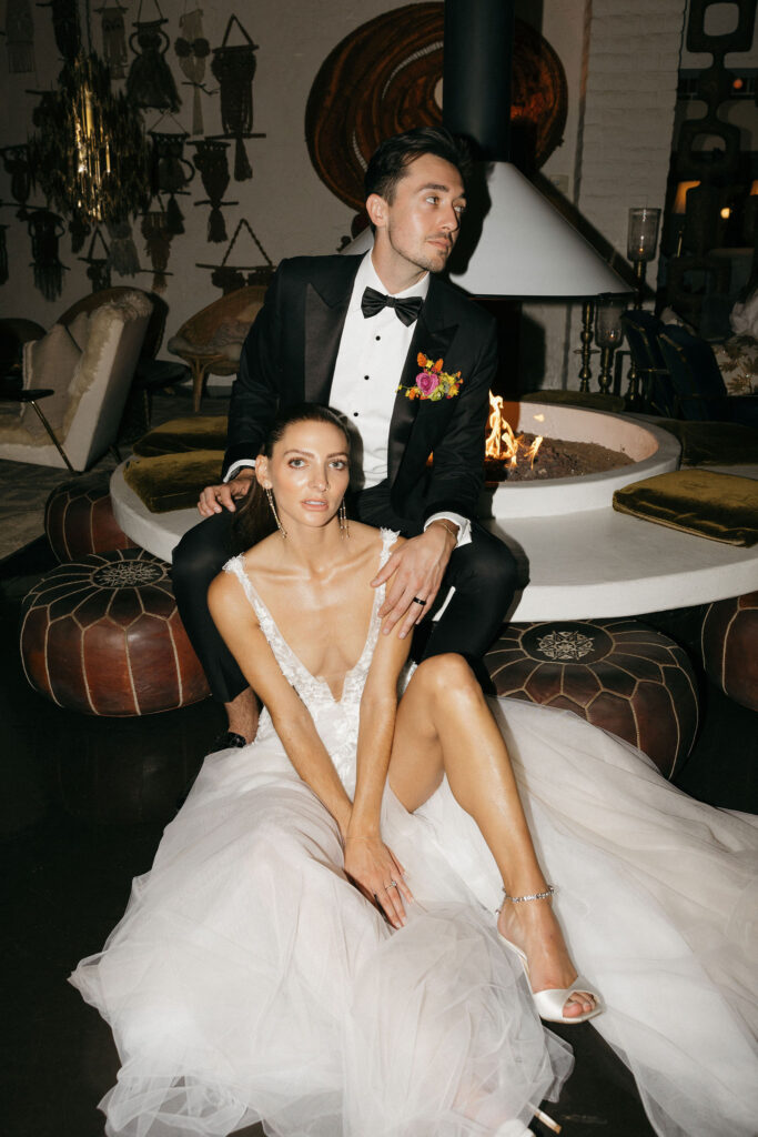 The bride sits gracefully in her wedding dress, leaning against the groom, who is seated on the edge of a fireplace in an indoor lounge area at Park Palm Springs Hotel, creating an intimate and stylish ambiance.