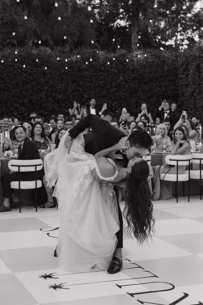 In a black-and-white photo, the groom dips the bride during their first dance, surrounded by guests capturing the moment on their phones at Park Palm Springs Hotel.