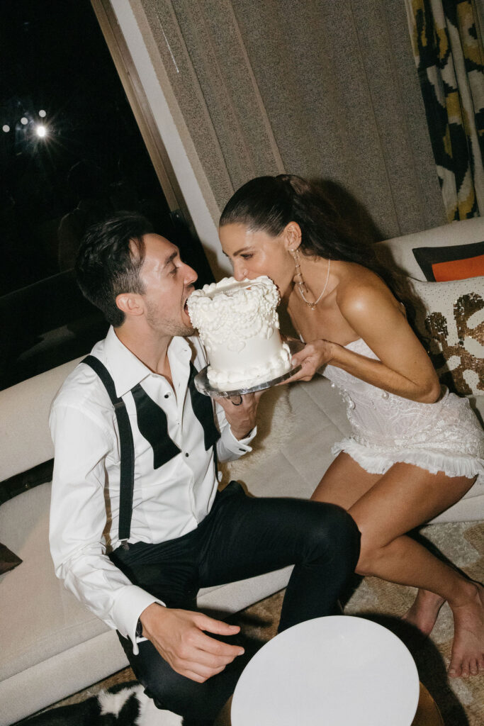 The bride and groom share a joyful moment as they feed each other cake, seated with a small, decorated cake on the table at Park Palm Springs Hotel.