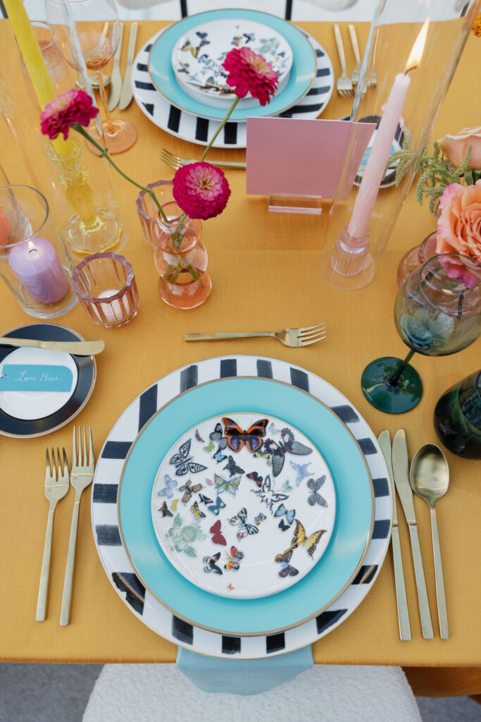 Vibrant wedding dinner table setting with pink, orange, and yellow decor - An elegant dinner table setting with pink and orange candles, bright flowers, and personalized name cards on butterfly-themed plates set atop a mustard yellow tablecloth. 