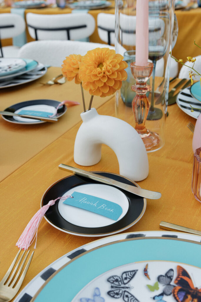 Place setting with a butterfly plate and vibrant orange dahlia in a white vase - A colorful and modern place setting featuring a butterfly motif plate, a unique white vase with an orange dahlia, and a pink tassel name card.