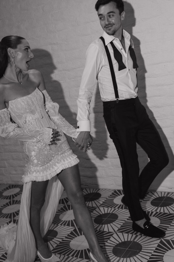 A black-and-white photo of the bride and groom dancing playfully, the bride wearing a short lace wedding dress with long sleeves and the groom in suspenders, at Park Palm Springs Hotel.