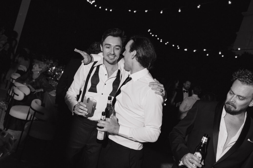 A joyful groom in a tuxedo, embracing friends and holding a drink at the reception.