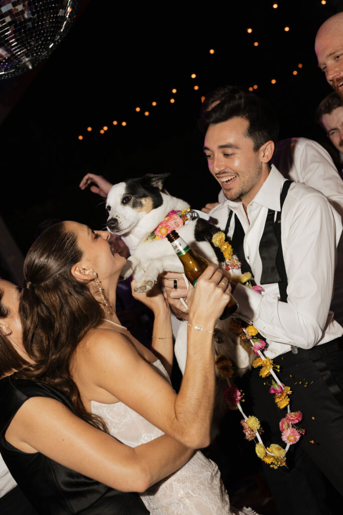 The bride and groom, smiling and holding their dog, surrounded by friends and a disco ball overhead at the reception.