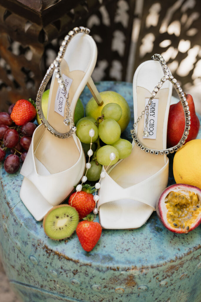 A pair of white Jimmy Choo high heels adorned with sparkling straps, placed next to a display of fresh fruit, including grapes, strawberries, and kiwi.