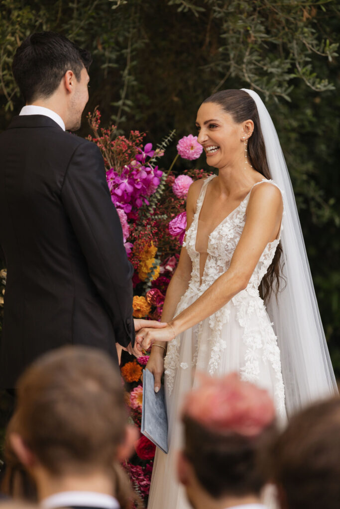 The bride, smiling brightly and holding the groom’s hands, standing in front of a floral arrangement during their ceremony.