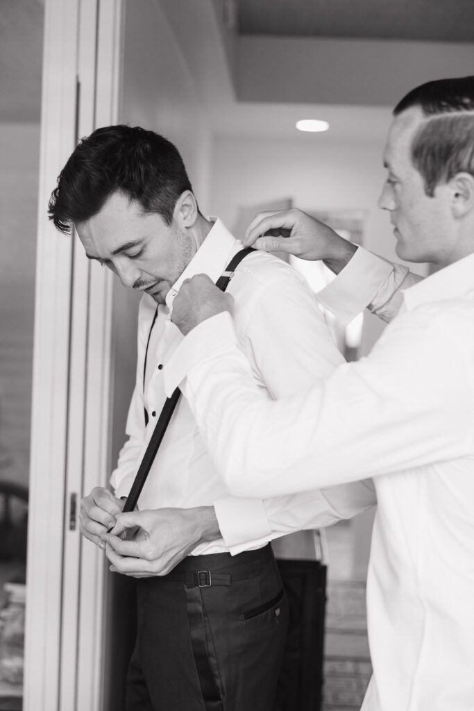 The groom getting ready at Park Palm Springs Hotel, assisted by a friend who adjusts his suspenders, in a candid black-and-white photo capturing the anticipation of the moment.