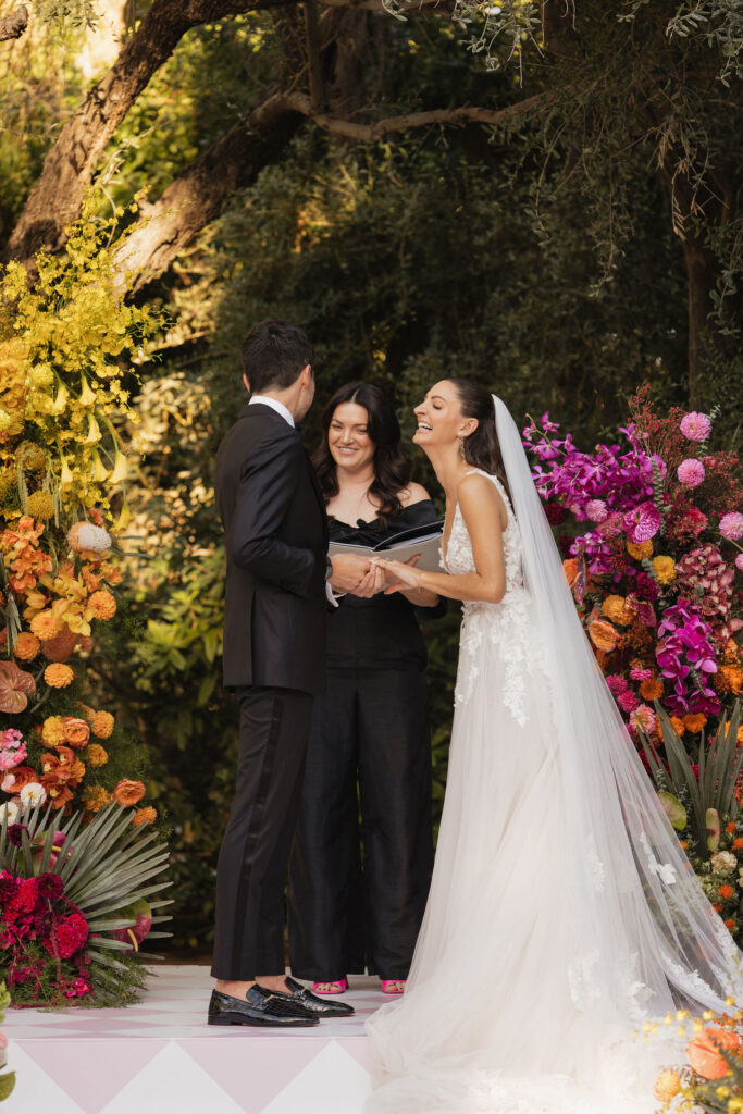 Wedding ceremony with officiant - The bride and groom share a light-hearted moment with their officiant, surrounded by vibrant floral arrangements in warm hues, at their outdoor wedding ceremony.