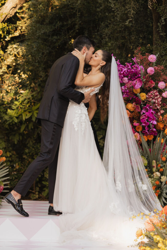 Bride and groom sharing a kiss at the altar - The bride and groom kiss passionately at the altar, surrounded by a vibrant floral display in pink, purple, and orange hues.