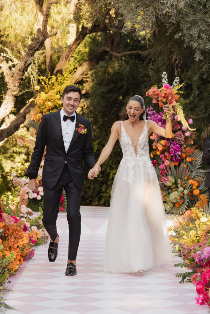 The bride and groom walk hand-in-hand down a flower-lined aisle, both beaming with joy, with the bride holding a vibrant bouquet of orange, pink, and yellow flowers.