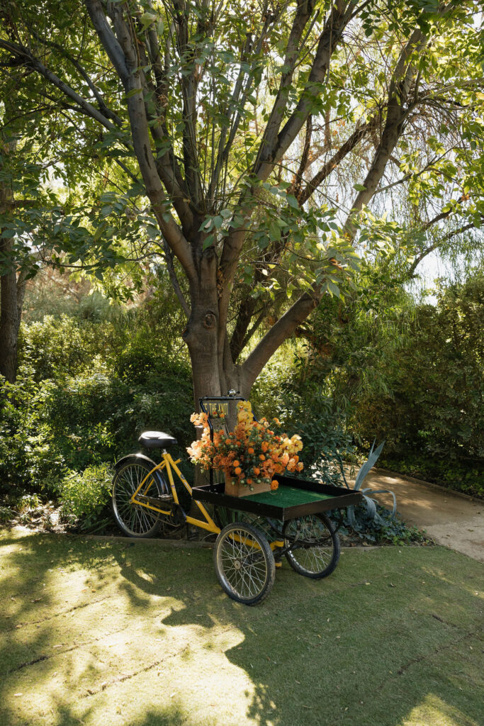 A vintage yellow bicycle with a basket of orange flowers is displayed under a tree in the Park Palm Springs Hotel garden.