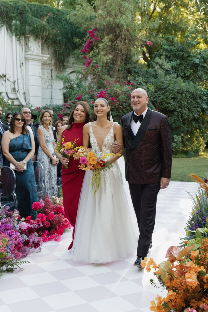 The bride, wearing a white dress and holding a vibrant bouquet, walks down the aisle accompanied by her parents in an outdoor ceremony.