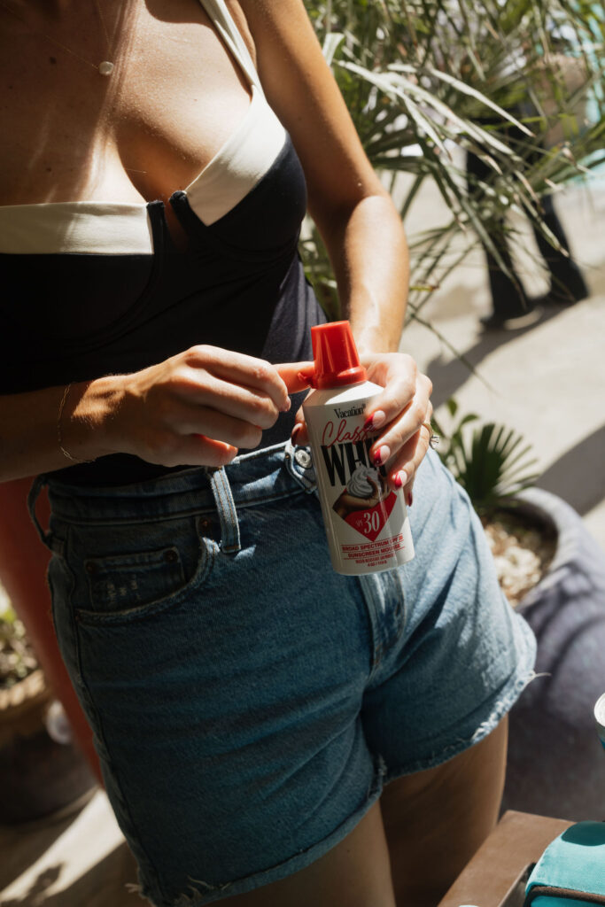 A guest applies sunscreen spray, wearing a black-and-white swimsuit and denim shorts, embracing the sunny weather at Park Palm Springs Hotel. 