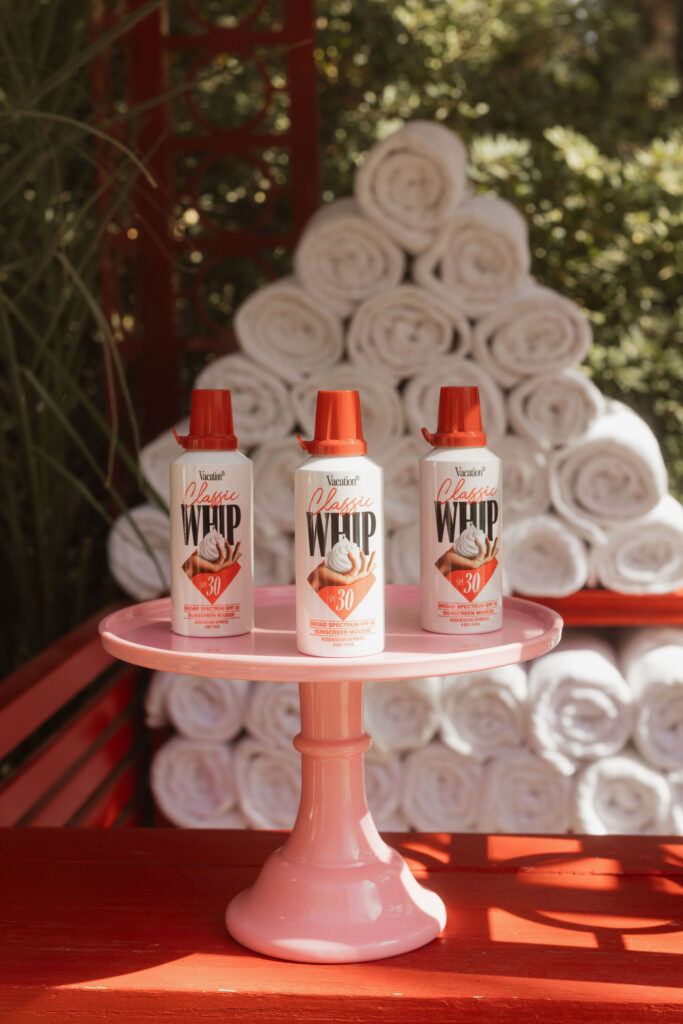 Three bottles of sunscreen mousse displayed on a pink cake stand in front of rolled white towels.