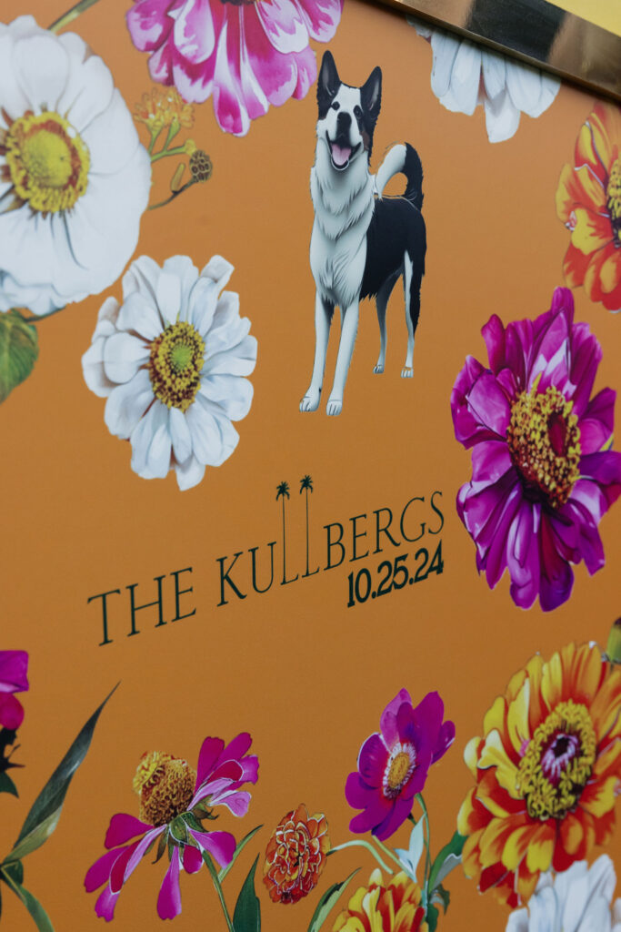 A vibrant orange sign decorated with colorful flowers and an illustration of a dog, reading "The Kullbergs 10.25.24."