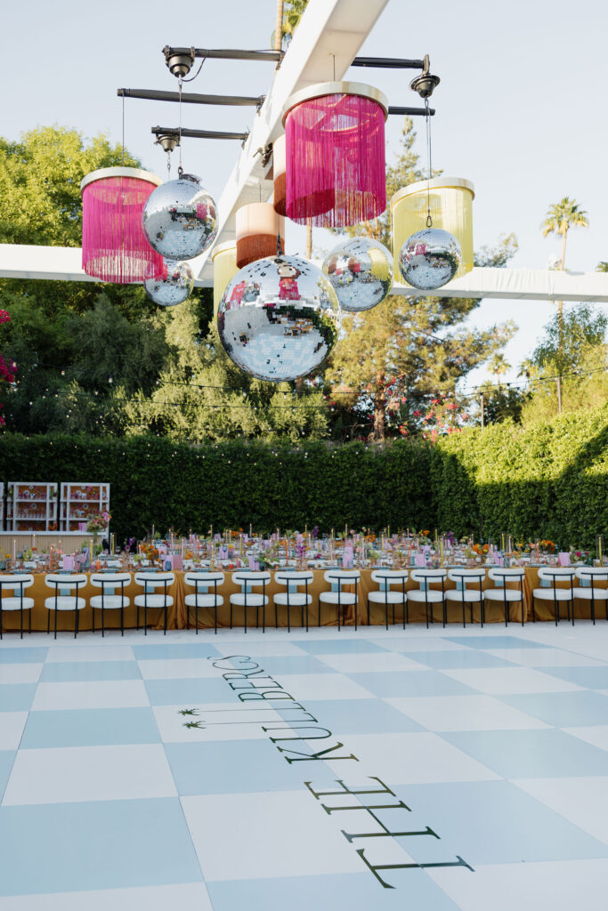 Rows of beautifully decorated tables with orange tablecloths, colorful glassware, and vibrant floral centerpieces, under a canopy with hanging disco balls and fringed lanterns at Park Palm Springs Hotel.