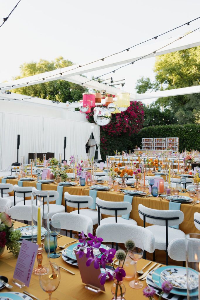 Rows of beautifully decorated tables with orange tablecloths, colorful glassware, and vibrant floral centerpieces, under a canopy with hanging disco balls and fringed lanterns at Park Palm Springs Hotel.