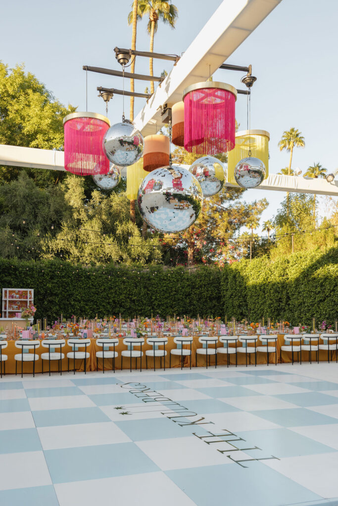 An elegant reception area with disco balls and colorful fringe decorations hanging above a long dinner table. The dance floor is personalized with "The Kullbergs" in bold letters, adding a chic touch to the outdoor event at Park Palm Springs Hotel.