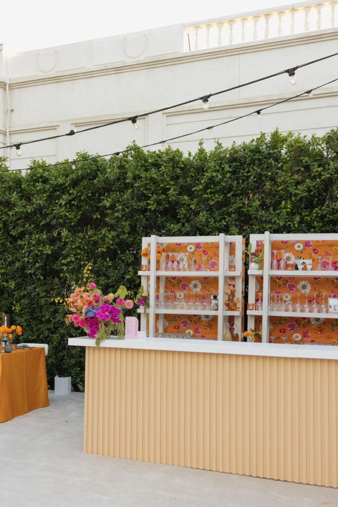 A beautifully decorated outdoor bar with a floral backdrop, colorful flowers, and lights strung overhead.