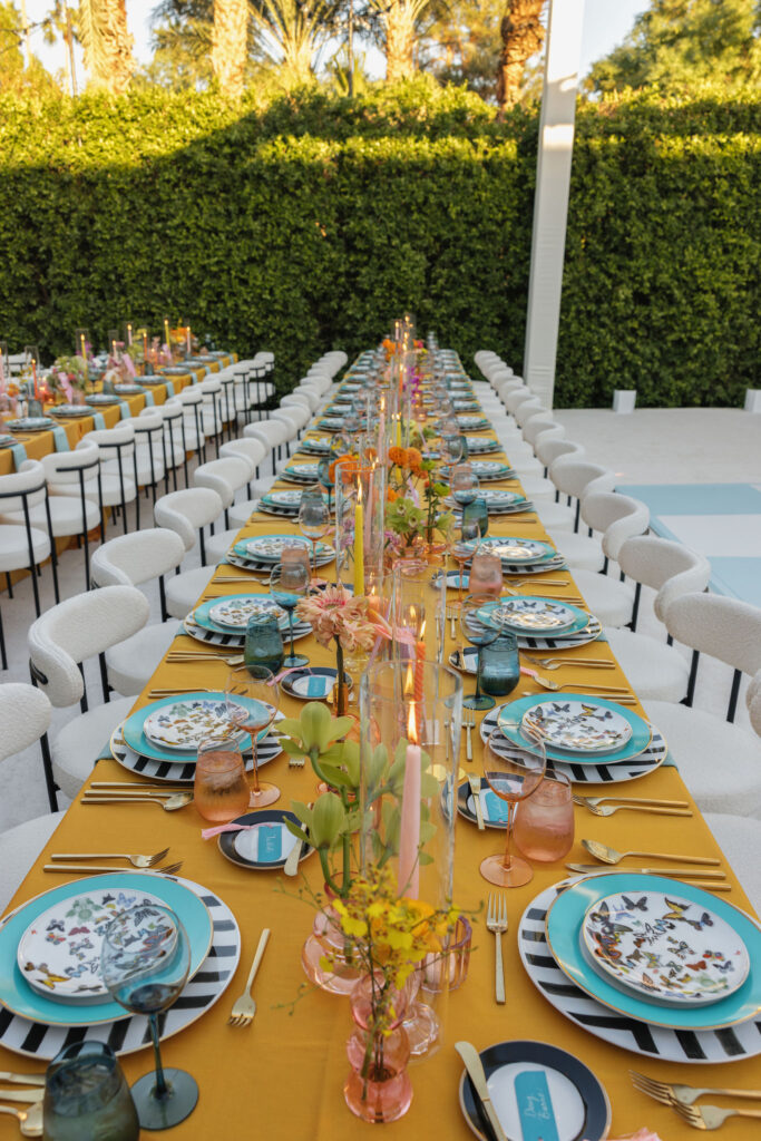 Long dining table with colorful decor and butterfly-themed plates - A long dining table setup for a wedding reception, decorated with butterfly plates, orange and pink candles, and lush green foliage in the background.