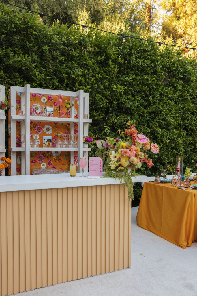 A wedding cocktail bar setup with a bold floral backdrop, displaying bright flower arrangements, and pink and yellow drinks on a modern bar counter.